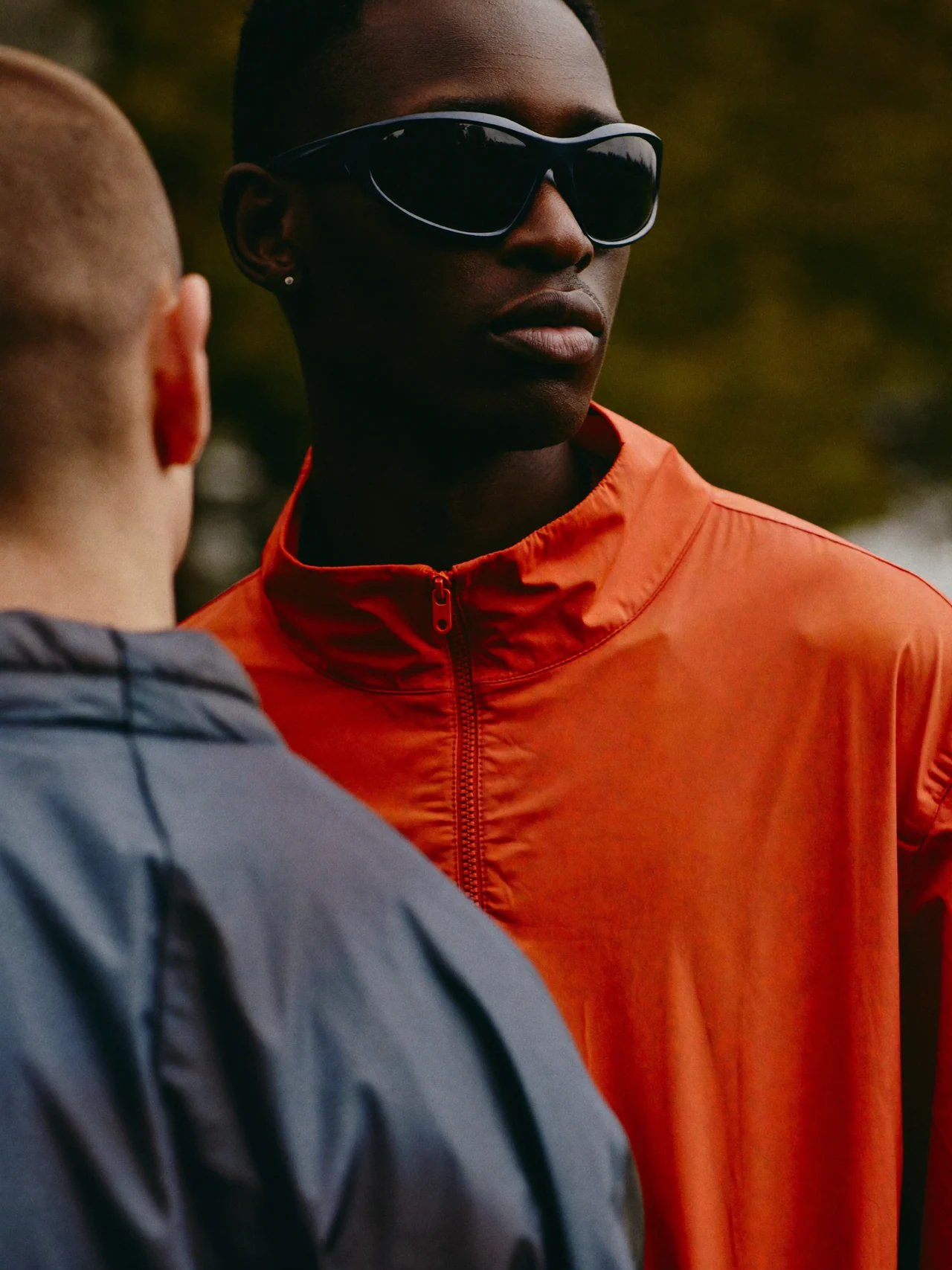 Orange technical outerwear and black sunglasses, framed by a figure in slate grey